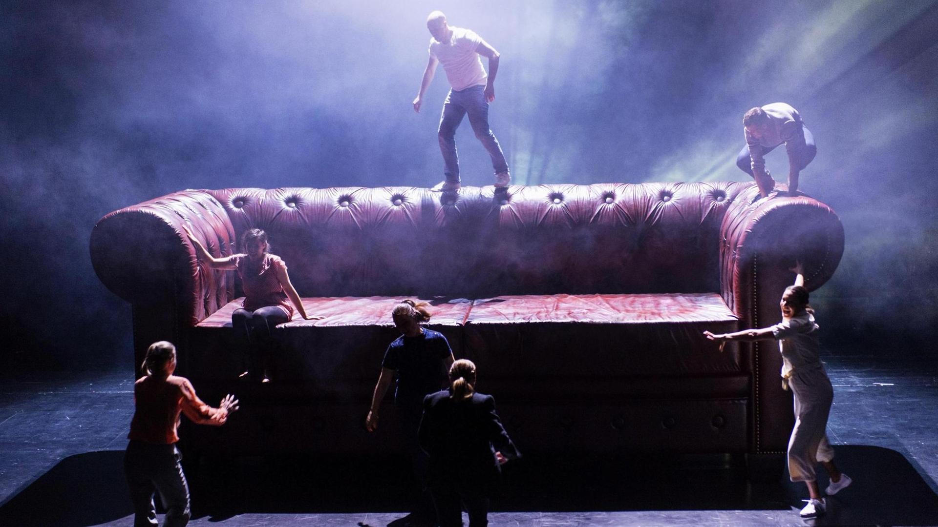 Eine Theater-Szene der Produktion HOUSE OF HORROR (Text von Volker Lösch und Christine Lang), die am Theater Bonn uraufgeführt wird. Das Zu sehen ist ein riesiges Ledersofa vor und auf dem sich Mitglieder des Ensembles bewegen.