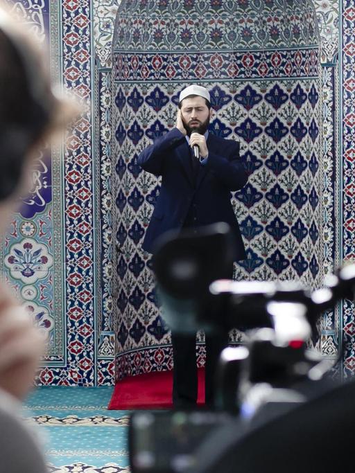 Ein Imam singt vor einer Kachelfliesenwand in rot und blau während er gefilmt filmt.