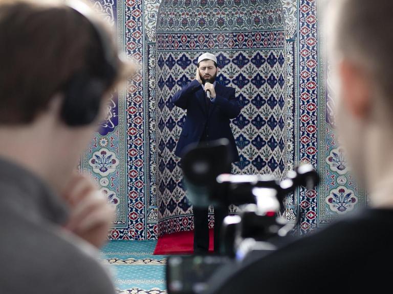 Ein Imam singt vor einer Kachelfliesenwand in rot und blau während er gefilmt filmt.