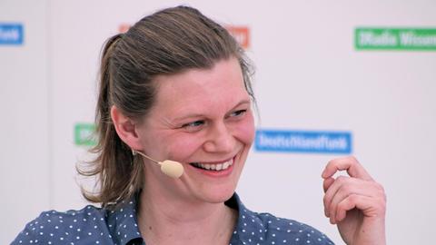 Eine Frau mit nach hinten gebundenen Haaren und einem Mikrofon schaut nach rechts aus dem Bild und lacht dabei.