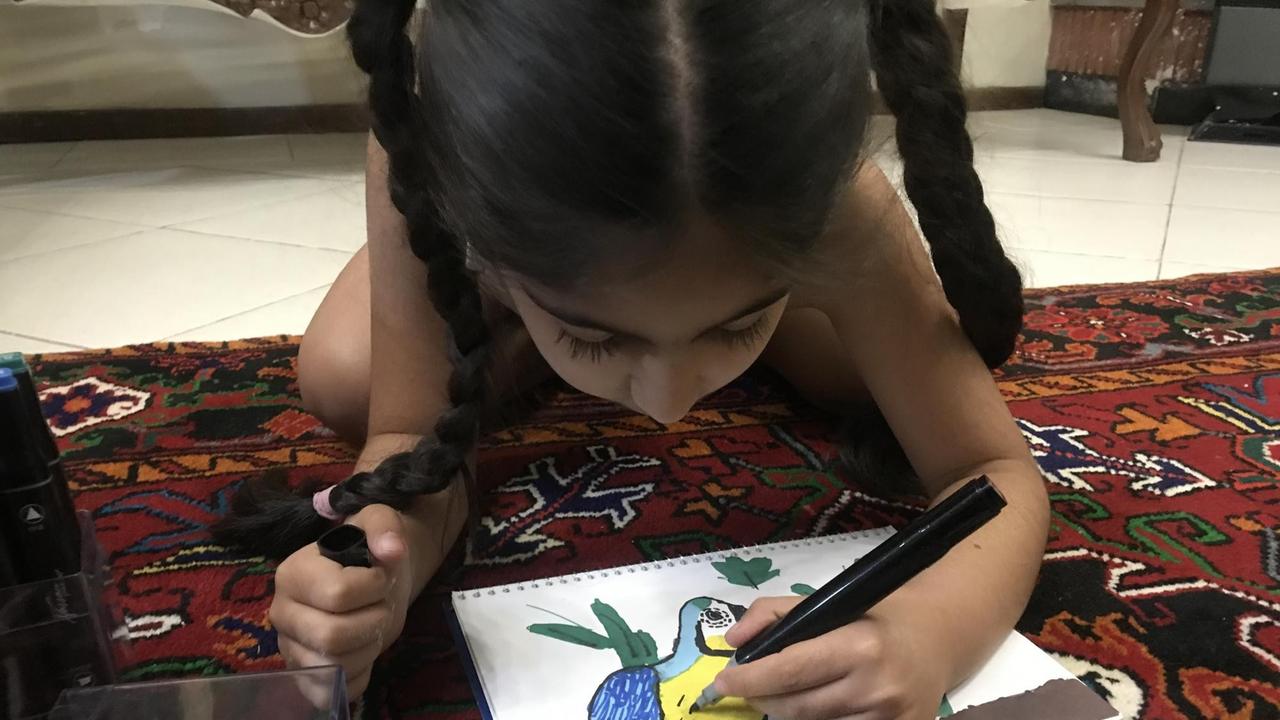 Ein kleines Mädchen mit zwei dunklen Zöpfen liegt auf einem Teppich und zeichnet einen Vogel.