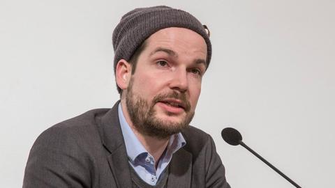 Max Czollek mit Hemd, Pullover und Mütze vor einem Mikrofon auf einer Bühne.
