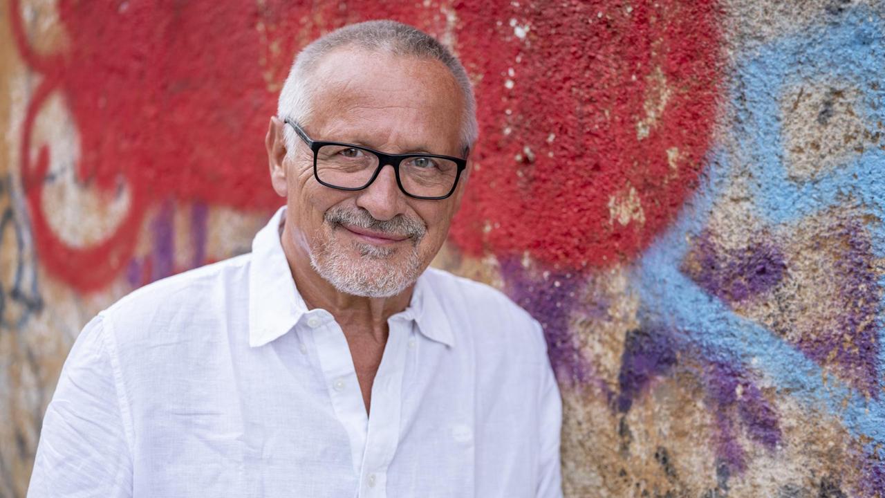Ein Mann in weißem Hemd, kurzem weiß-grauen Haar und Bart lächelt in die Kamer. Er ist im Halbporträt vor einer bunt bemalten Wand zu sehen.