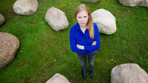 Vogelperspektive einer blonden Frau, die mit verschränkten Armen zwischen großen Steinen auf einer Wiese steht.