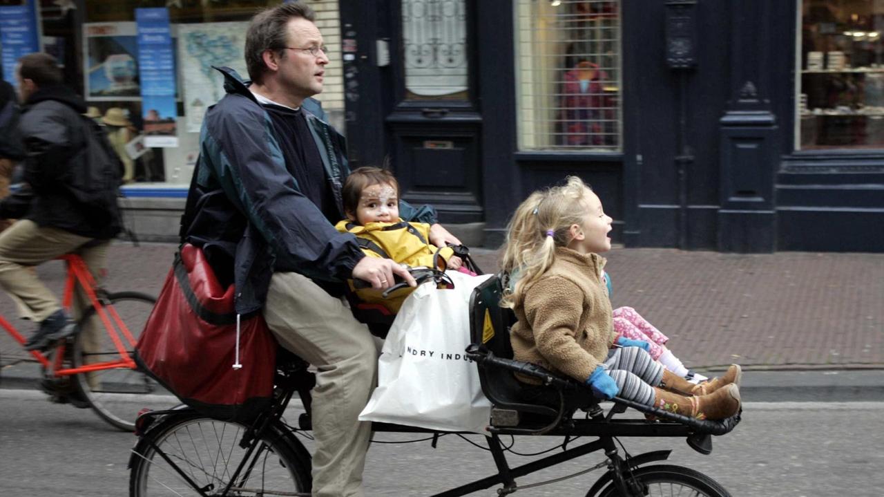 Auf einem ungewöhnlichen Zweirad radelt ein Mann mit seinen beiden Kindern, die in Kindersitzen vorne sitzen, durch die Innenstadt von Amsterdam.