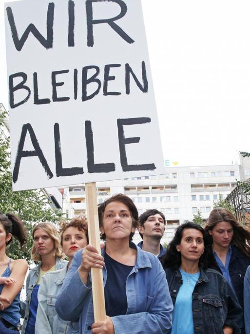 Die Autorin Christiane Rösinger ist mit ihrem Cast zu sehen, sie hält ein Schild mit der Aufschrift "Wir bleiben Alle" hoch.