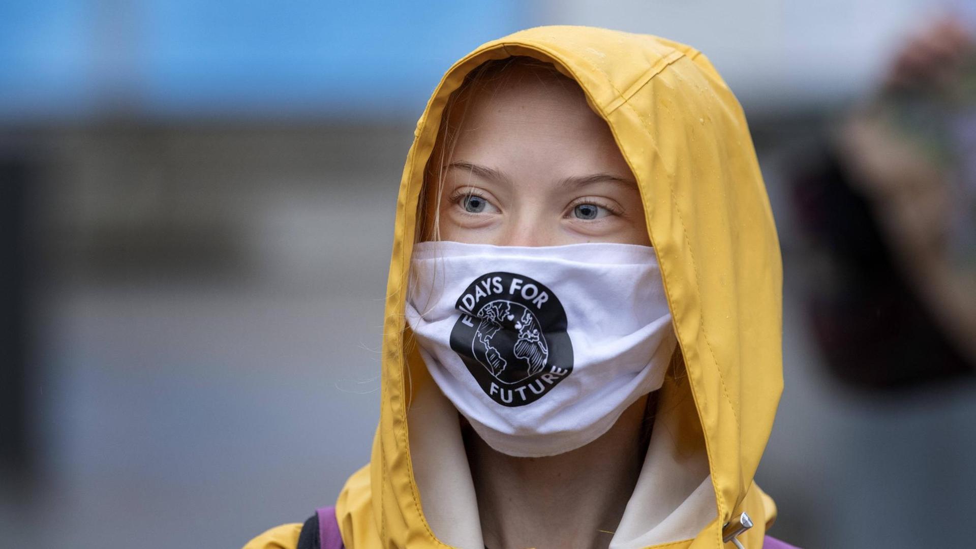 Porträt der schwedischen Klimaaktivistin Greta Thunberg mit Mund-Nasen-Schutz und gelber Regenmantel-Kapuze.