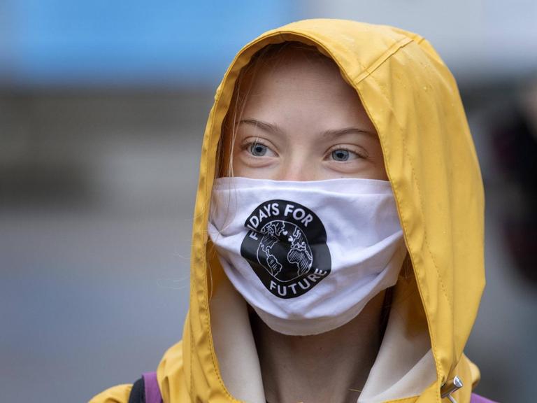 Porträt der schwedischen Klimaaktivistin Greta Thunberg mit Mund-Nasen-Schutz und gelber Regenmantel-Kapuze.