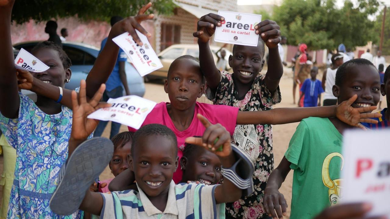 Kinder und Jugendliche haben Zettel in der Hand mit der Aufforderung einen Wahlschein zu holen. Die senegalesische Bevölkerung ist sehr jung, im Schnitt 18 Jahre.