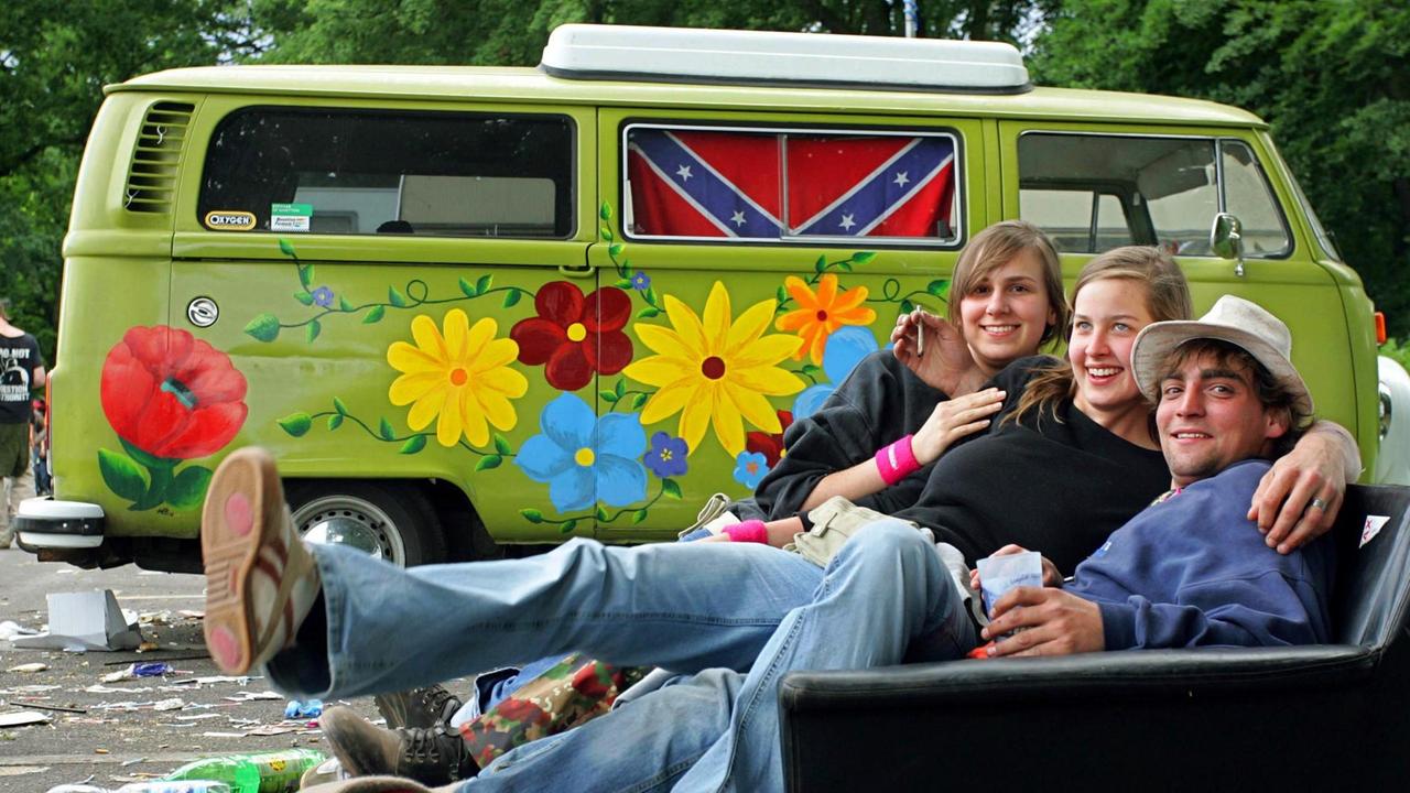 Drei Besucher des Festivals "Rock im Park" sitzen auf einem Campingplatz, vor einem VW-Bulli, auf einer alten Couch. 