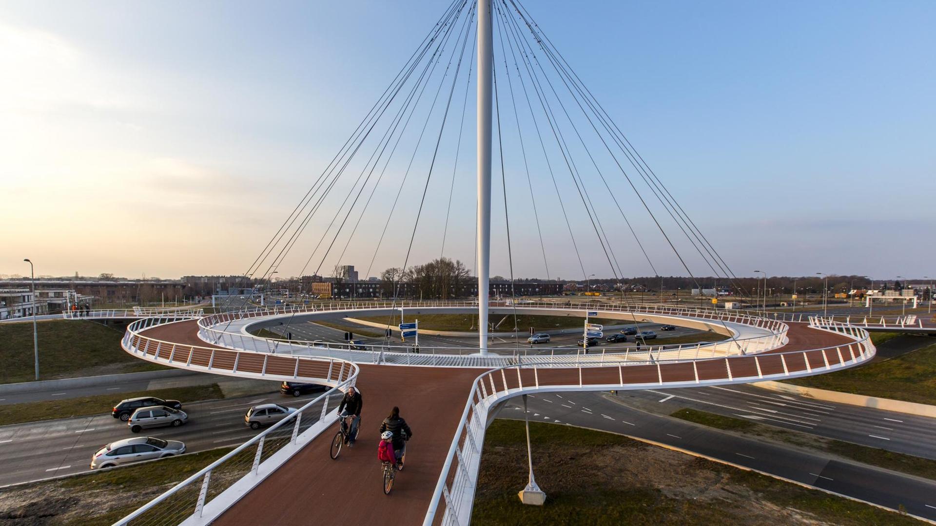 Ein futuristischer Bau in Form eines Kreisels für Fahrradfahrer schwebt über einer Autostraßenkreuzung.