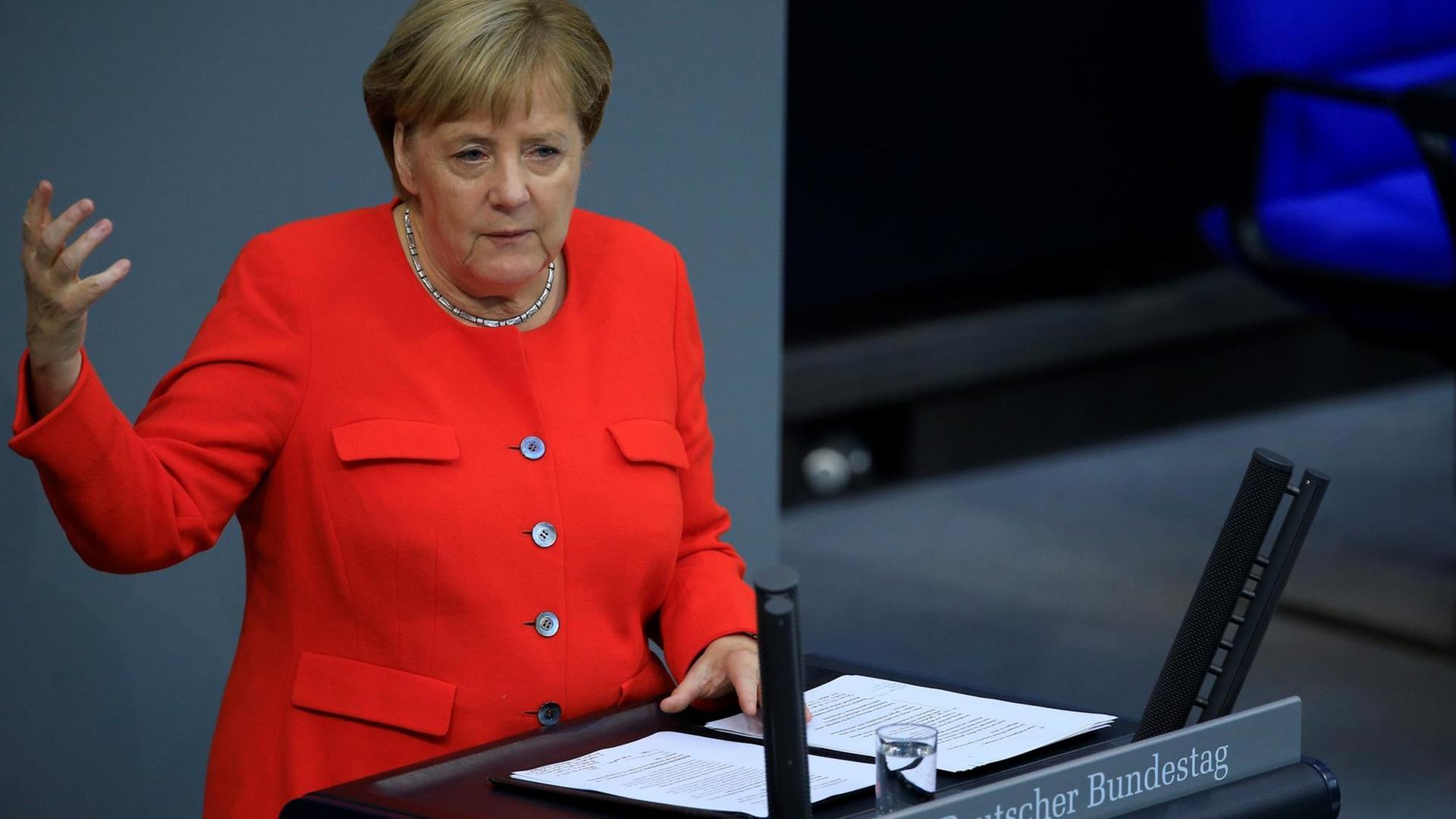 Angela Merkel spricht am Rednerpult des Deutschen Bundestages. Sie trägt eine rote Jacke, die linke Hand berührt das Rednerpult, die rechte hält sie angewinkelt bis in Schulterhöhe in die Luft.