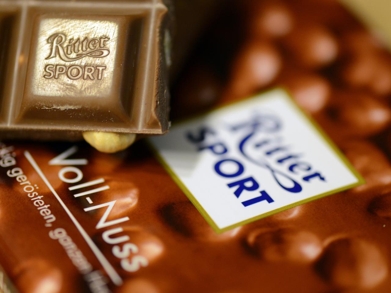 Das Bild zeigt ein kleines Stück Voll-Nuss-Schokolade der Marke Ritter Sport. Es liegt auf einer Verpackung der Schokoladentafel.