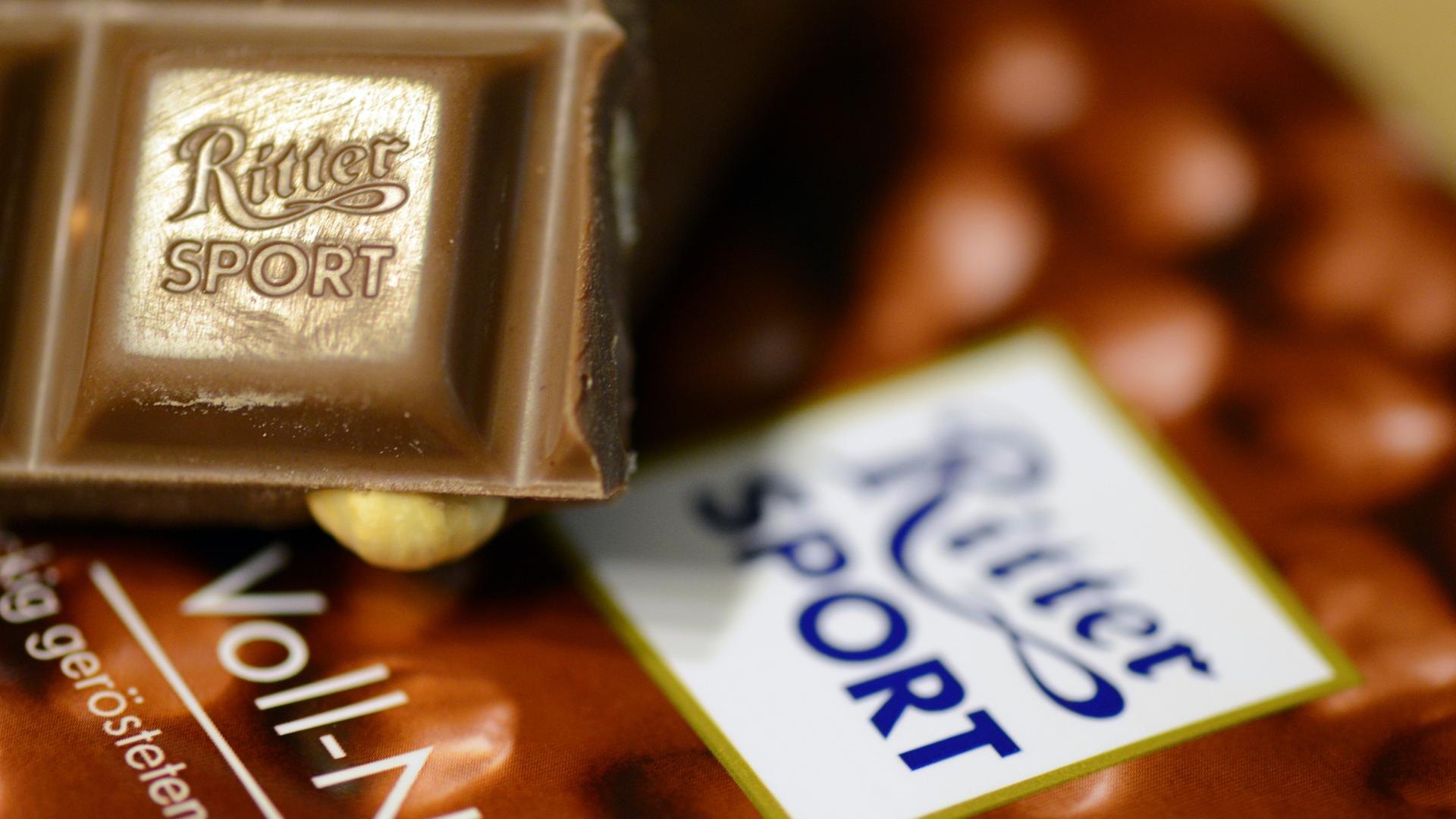 Das Bild zeigt ein kleines Stück Voll-Nuss-Schokolade der Marke Ritter Sport. Es liegt auf einer Verpackung der Schokoladentafel.