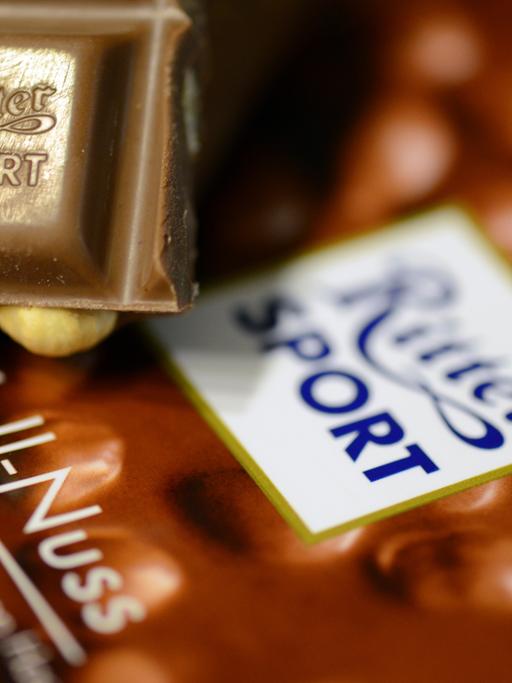 Das Bild zeigt ein kleines Stück Voll-Nuss-Schokolade der Marke Ritter Sport. Es liegt auf einer Verpackung der Schokoladentafel.
