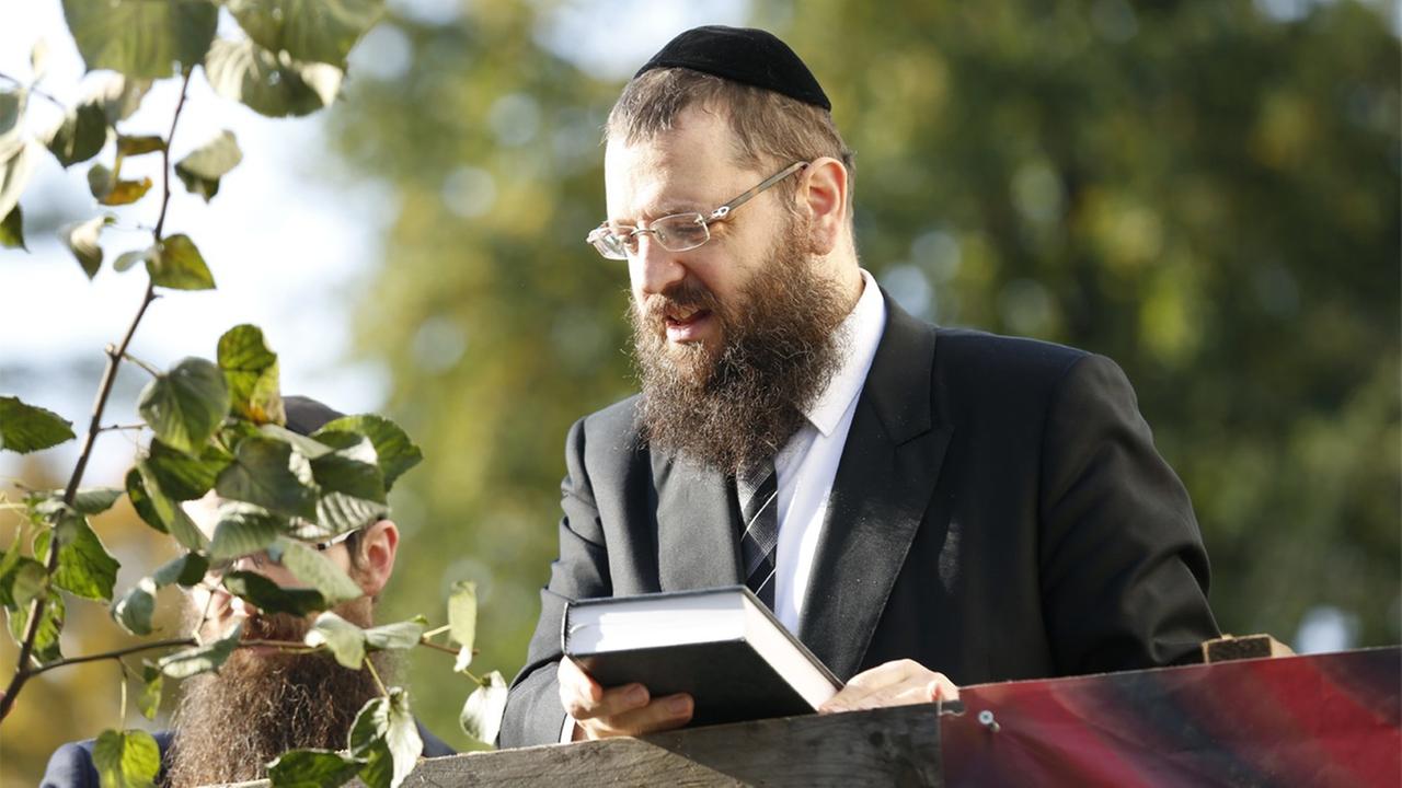 Ein Rabbiner steht auf einer Holzbühne und spricht zu Menschen.