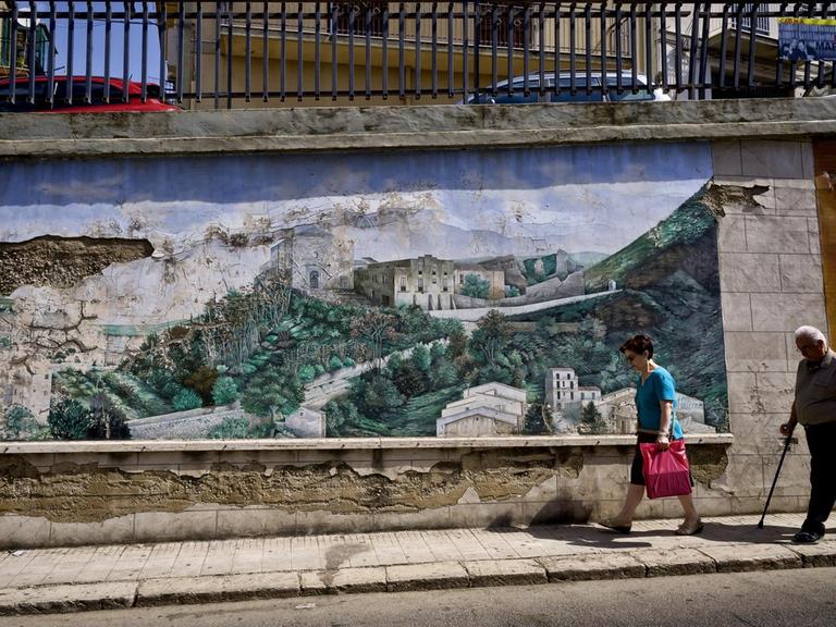 Eine Straßenstzene mit zwei älteren Passanten in Corleone, die an einem Wandbild mit dem Motiv einer ländlichen Landschat vorbeigehen.