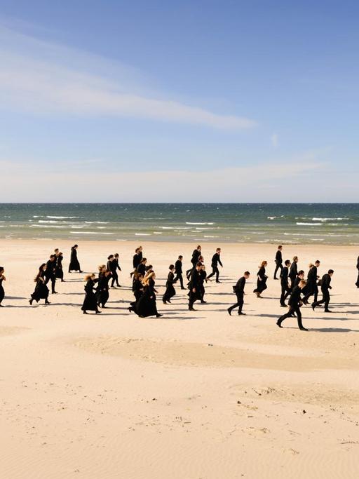 Die Orchestermitglieder laufen in schwarzer Kleidung einen langen Strand entlang.