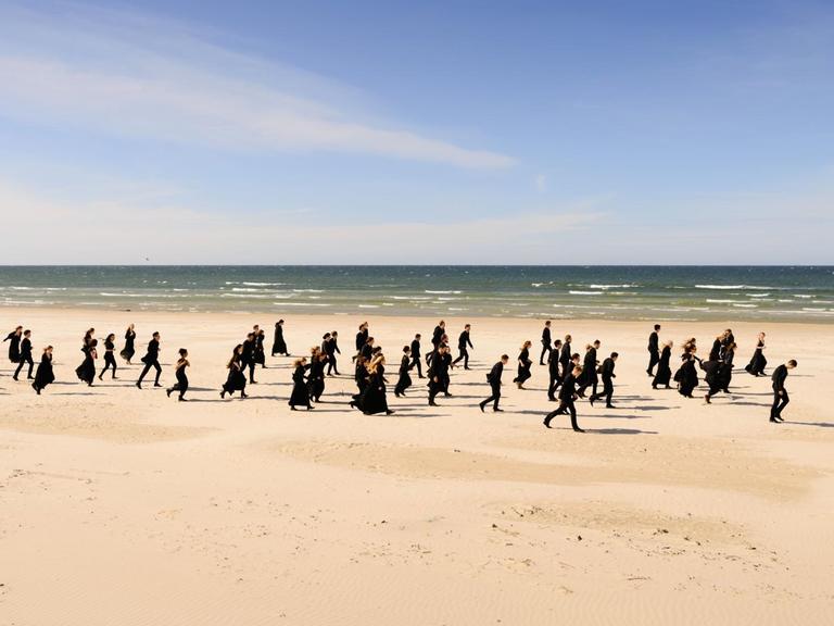 Die Orchestermitglieder laufen in schwarzer Kleidung einen langen Strand entlang.