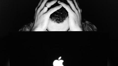 Ein Mann sitzt hinter einem Laptop und hat den Kopf in die Hände gestützt. 