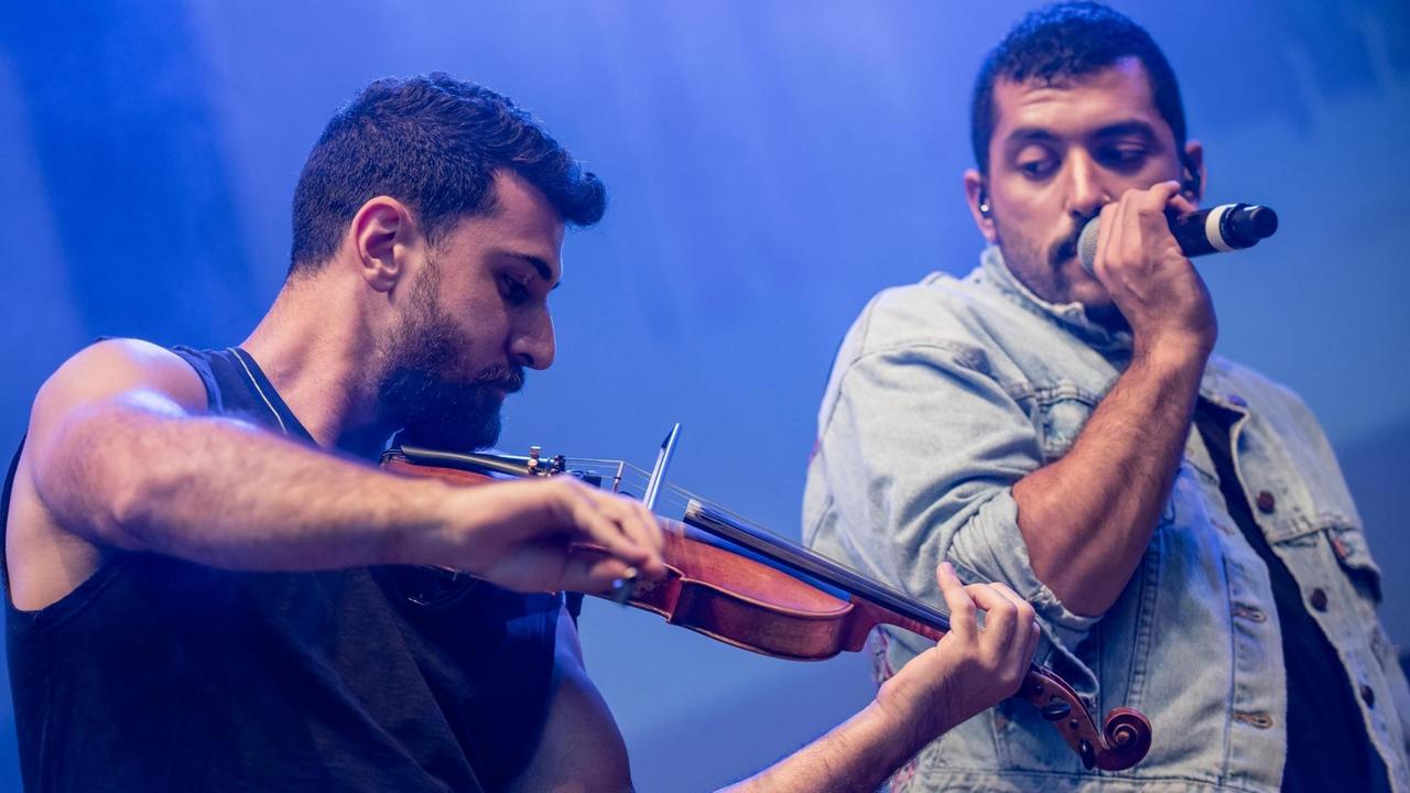 Zwei Männer stehen auf einer Bühne. Ein Mann spielt Geige, der zweite Mann singt in ein Mikrofon.