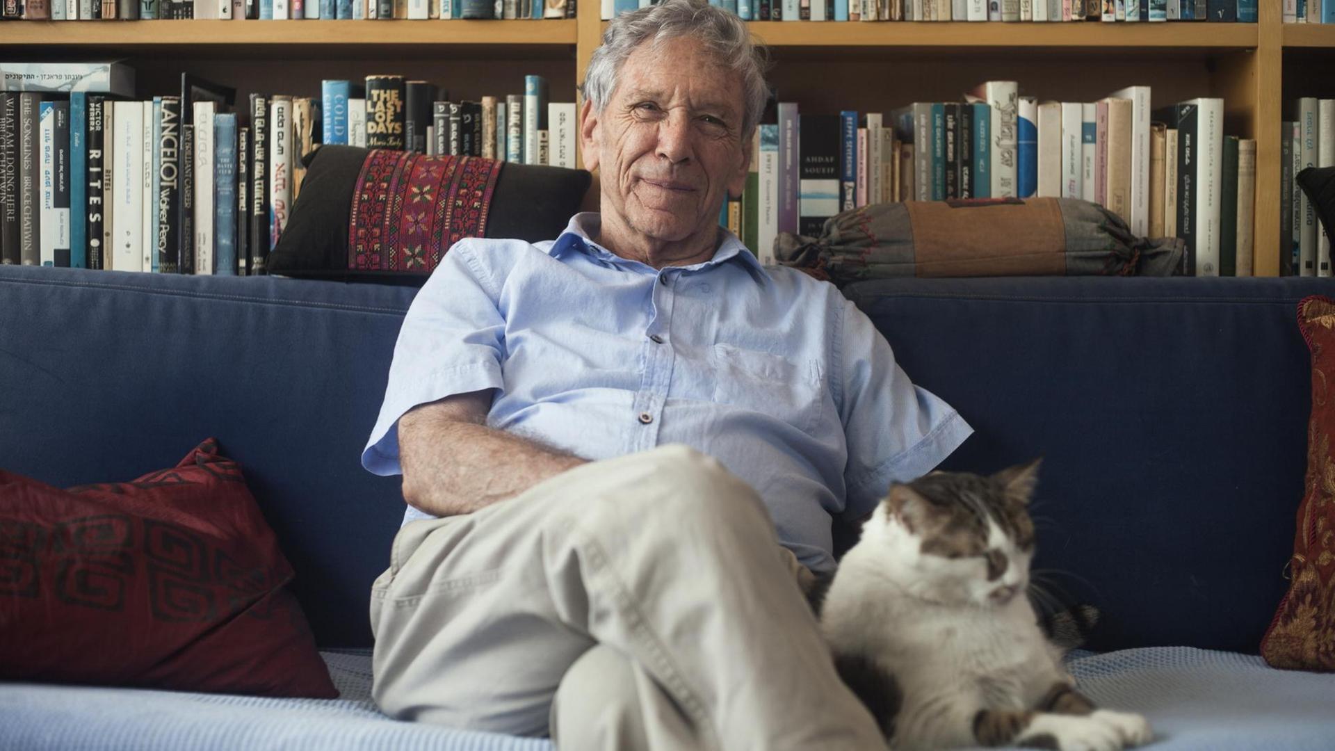 Der Schriftsteller Amos Oz sitzt auf einem Sofa. Hinter ihm eine Bücherwand. Neben ihm sitzt eine Katze.