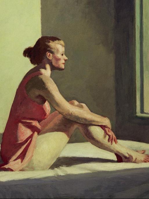 Eine junge Frau sitzt auf einem Bett und blickt melancholisch aus dem Fenster.