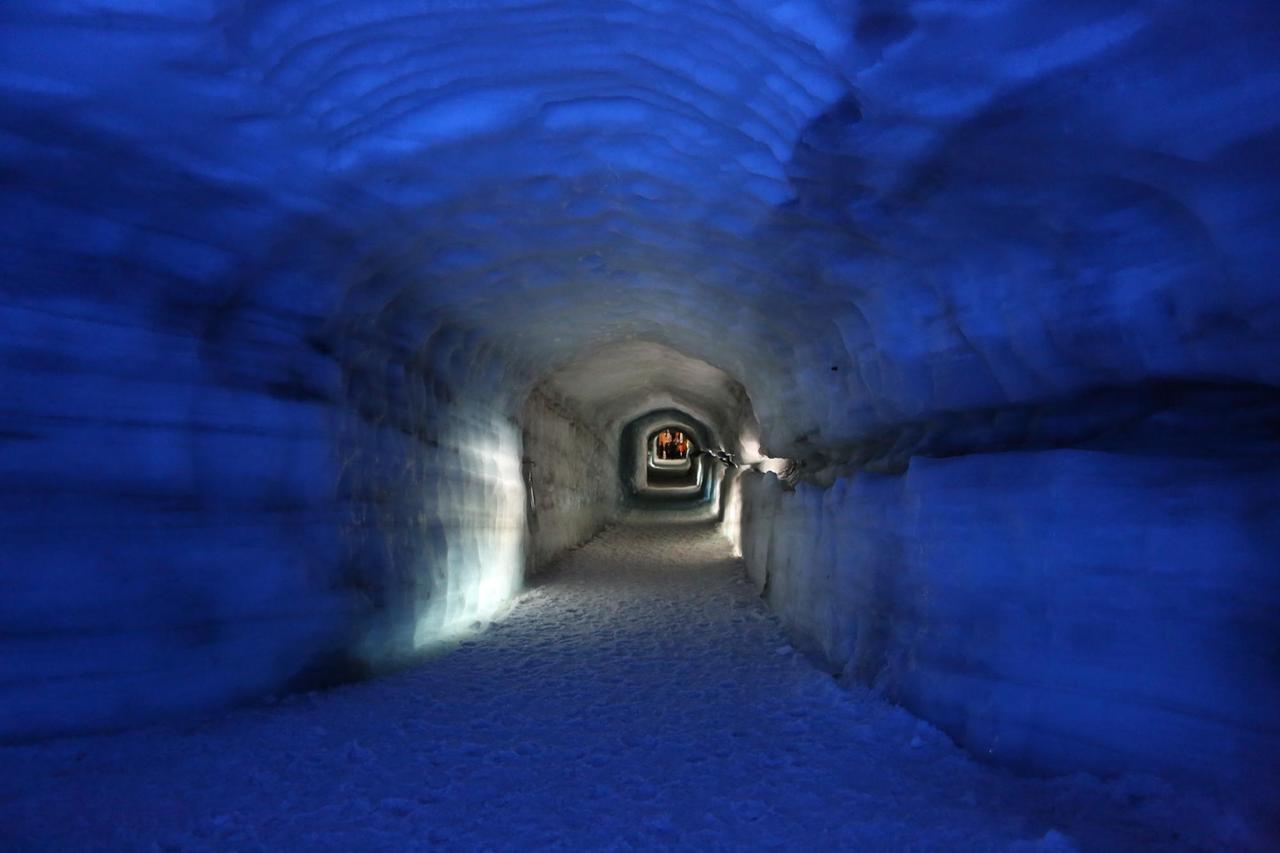 Das Eis des Tunnelgangs ist im Vordergrund in dunklem Blau angeleuchtet. Weiter hinten wird es weiß, ganz hintern ist eine Gruppe von menschen zu sehen.