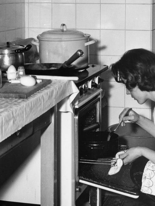 Undatierte Aufnahme aus den 1950er oder 1960er Jahren: Eine junge Hausfrau kniet vor einem Ofen und kontrolliert einen Braten.