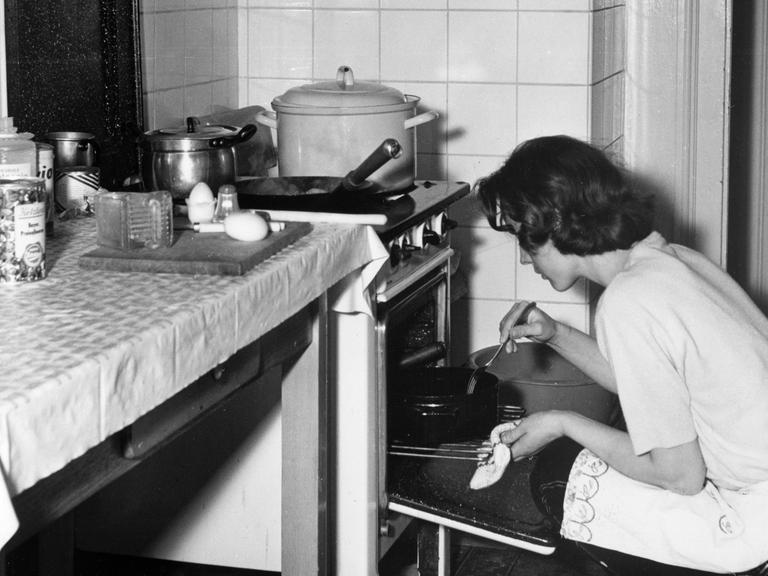 Undatierte Aufnahme aus den 1950er oder 1960er Jahren: Eine junge Hausfrau kniet vor einem Ofen und kontrolliert einen Braten.