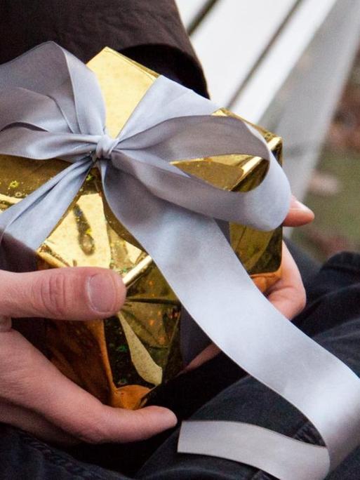 Ein junger Mann sitzt auf einer Bank und hält ein kleines, in goldenes Papier verpacktes, Geschenk in der Hand.