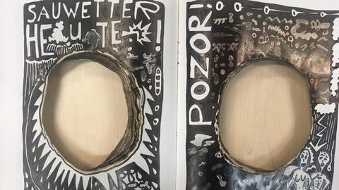 Kunstwerk in Form eines aufgeschlagenen Buches, beide Seiten mit einem Loch in der Mitte, links die Aufschrift "Sauwetter heute", rechts "Pozor"