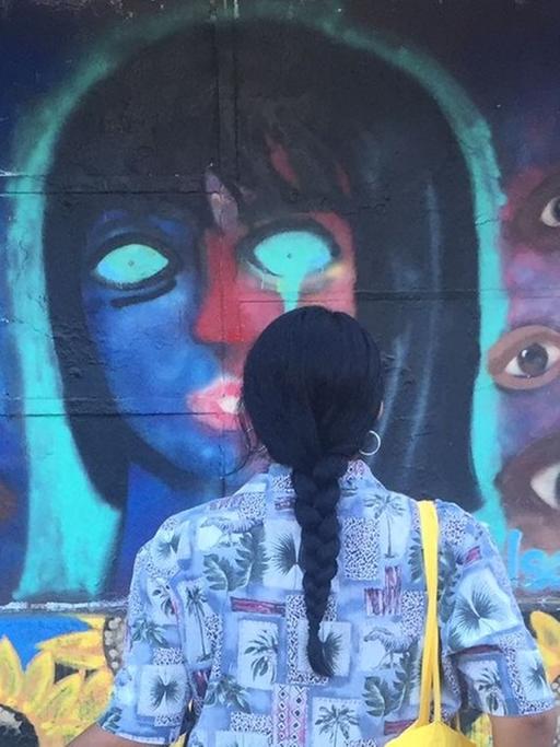 Eine junge Frau mit langem schwarzen Zopf und Fahrrad betrachtet ein Wandgemälde, das ein Frauengesicht mit leeren Augenhöhlen zeigt, drumherum viele Augen.
