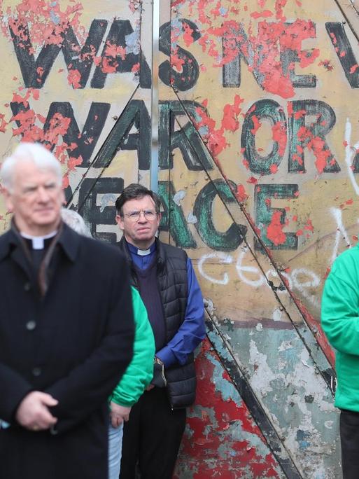 Vor einer Mauer mit dem Spruch "There was never a good war or a bad peace" stehen Menschen mit gefalteten Händen.