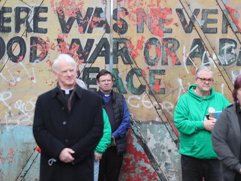 Vor einer Mauer mit dem Spruch "There was never a good war or a bad peace" stehen Menschen mit gefalteten Händen.