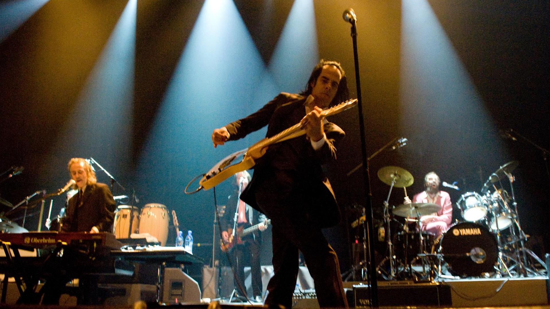 Ein Mann mit Gitarre in der Hand steht auf einer Bühne, im Hintergrund weitere Musiker.
