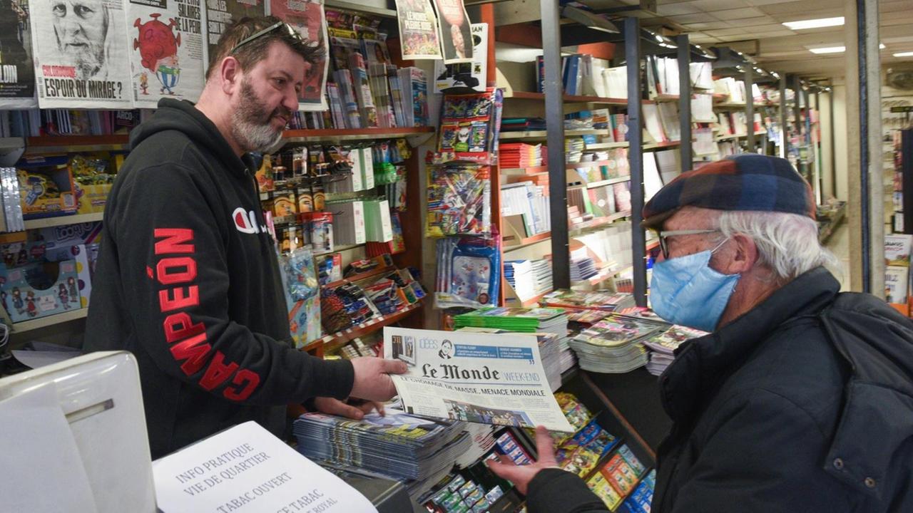 Ein Kioskbesitzer in Paris verkauft einem Kunden mit Mundschutz eine Tageszeitung