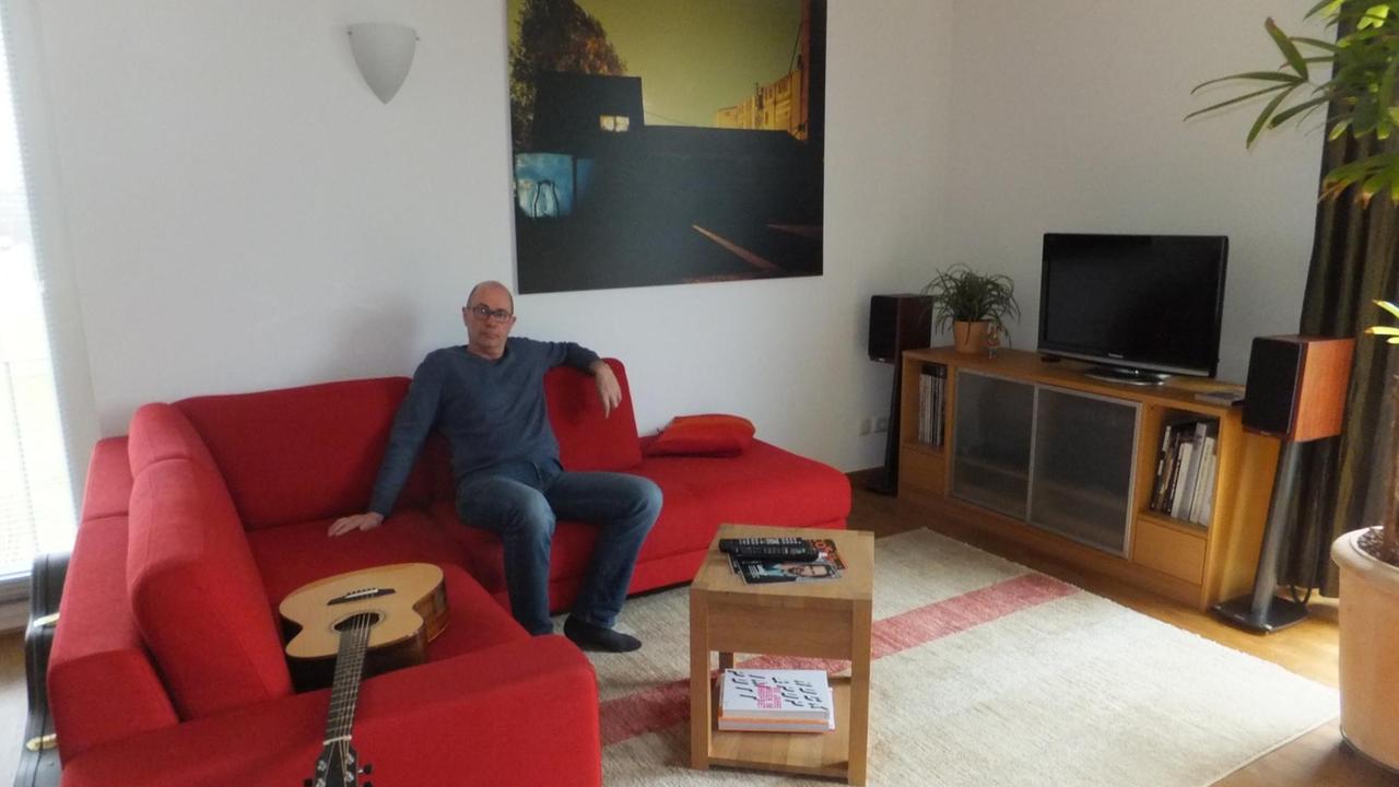 Uwe Lyko sitzt in seinem Wohnzimmer auf einem großen roten Sofa, auf dem auch eine Gitarre liegt