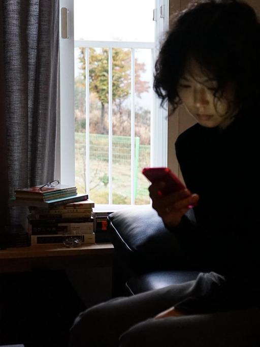 Filmstill aus "The Woman Who Ran": Eine Frau sitzt, auf das Smartphone blickend, vor einem Fenster mit halb zugezogenen Vorhängen.