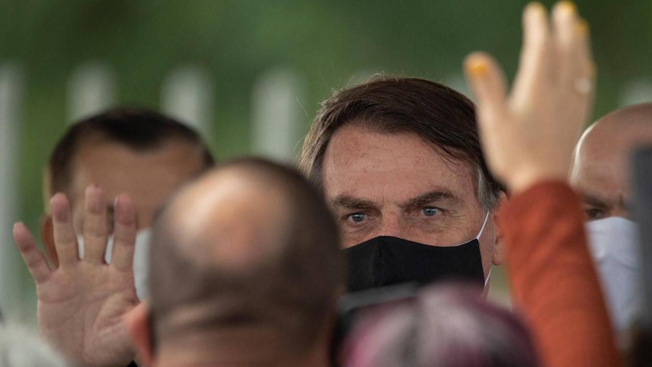 Großaufnahme von Präsident Bolsonaro, der einen schwarzen Mund-Nasenschutz trägt. Sein Gesicht wird halb verdeckt von Anhängern, die ihm zuwinken.