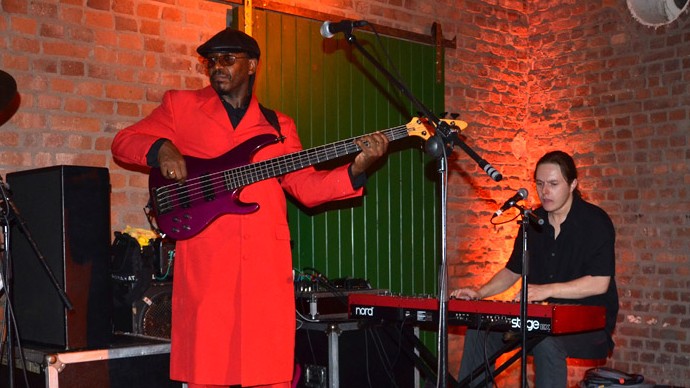 Ein Mann in rotem Anzug steht mit einer Gitarre auf der Bühne. Hinter ihm sitzt ein Mann am Schlagzeug.