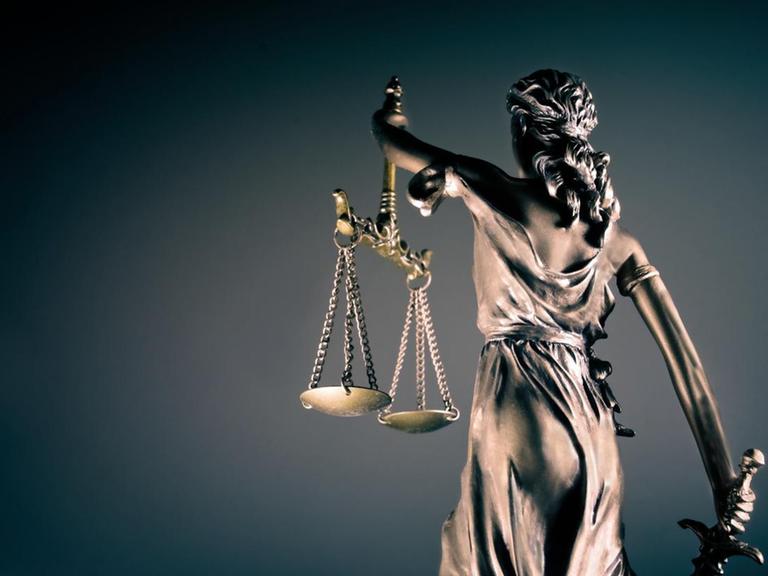 Vor einem dunklen Hintergrund steht dem Betrachter mit dem Rücken zugewandt die Figur der Justitia, die in der rechten Hand ein Schwert und in der linken Hand zwei Waageschalen trägt.