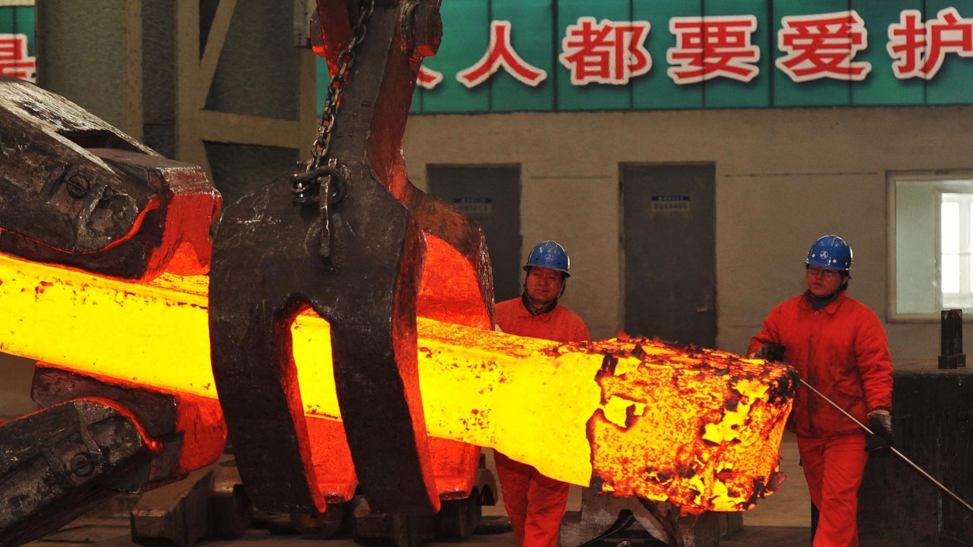 Eine rotglühender Stahlträger wird von einem Greifarm gehalten, zwei Arbeiter stehen in Schutzanzügen daneben