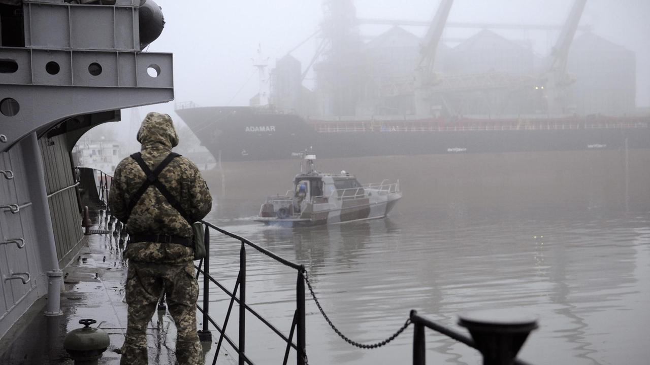 Ein ukrainischer Soldat auf einem Militärschiff im Hafen von Mariupol. Im Hintergrund fährt ein Patrouillenboot vorbei.