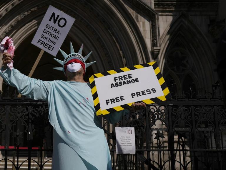 Ein als Freiheitsstatue verkleideter Mann hält ein Schild hoch, auf dem "Free Assange. Free Press" steht