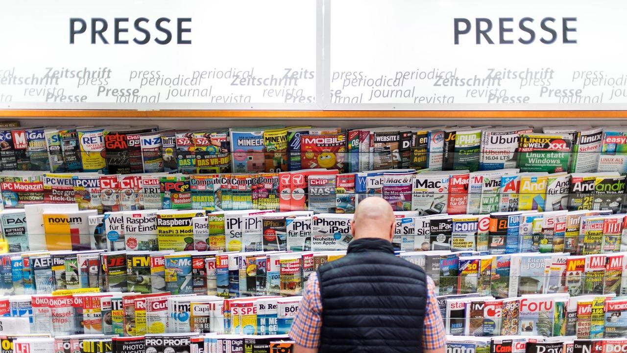 Der Mann mit Glatze ist von hinten zu sehen, vor ihm der Stand mit vielen Zeitschriften. Oben stehen die Worte "Presse" und "Zeitschrift" in mehreren Sprachen.