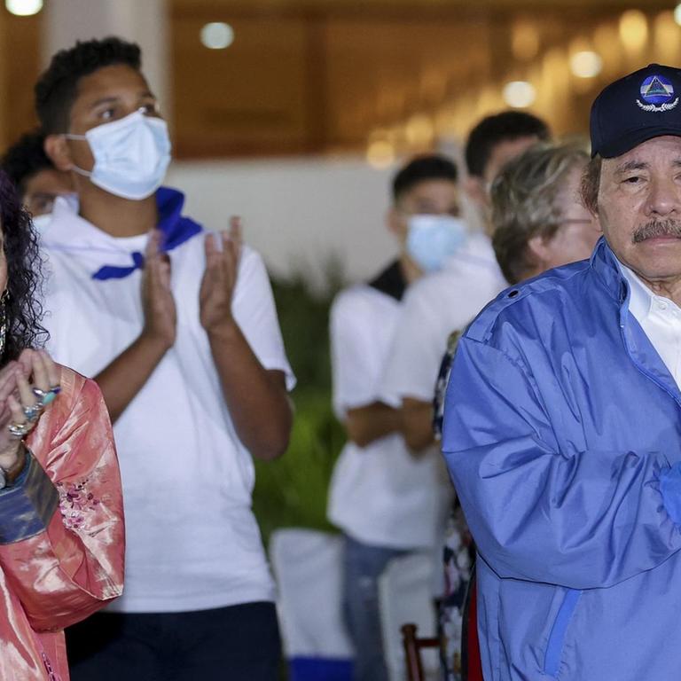 Auf dem Bild klatschen Daniel Ortega und Rosario Murillo in die Hände. Ortega trägt eine hellblaue Jacke und eine dunkle Kappe. Murillo at lange dunkle Locken und trägt ein rosa Kleid.
