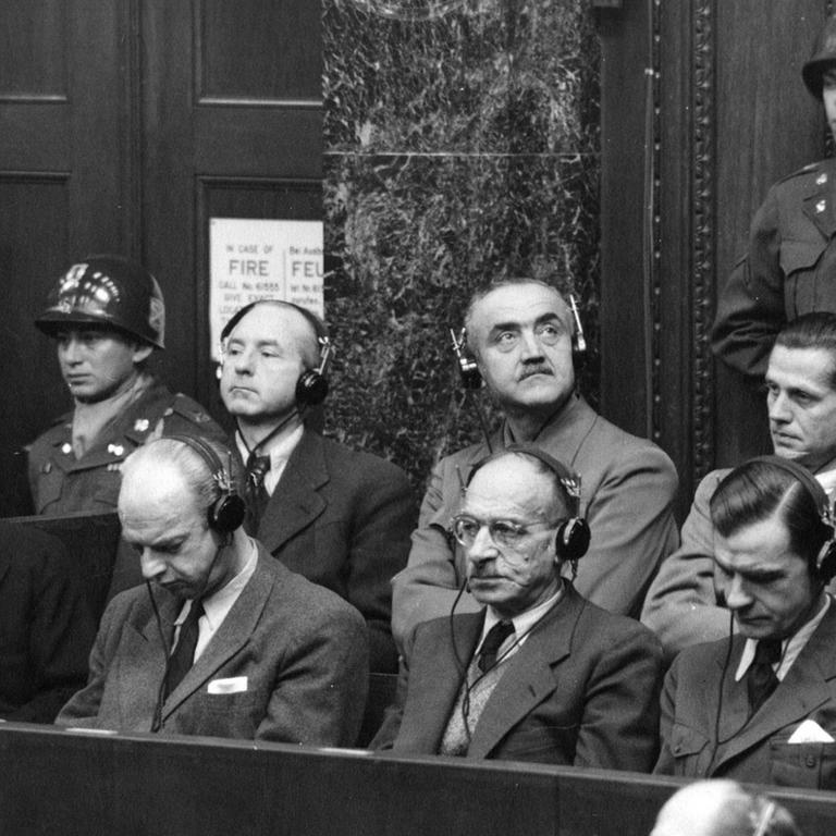 Männer in Anzügen sitzen auf einer Anklagebank ein einem Gerichtssaal.