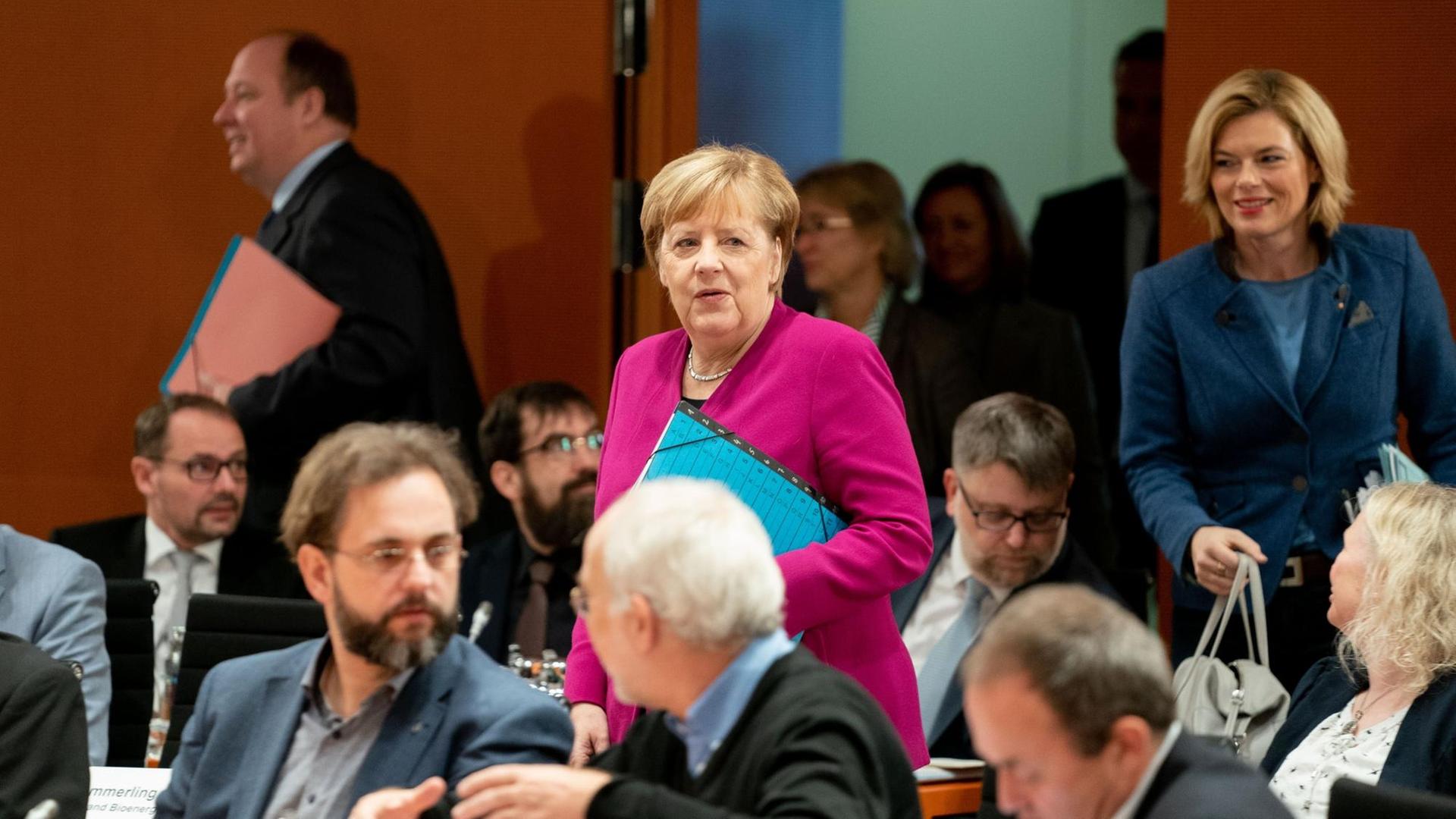 Merkel im roten Kostüm und Klöckner im blauen Kostüm mit Handtasche gehen gerade in den Saal rein, in dem schon zahlreiche Teilnehmer sitzen.