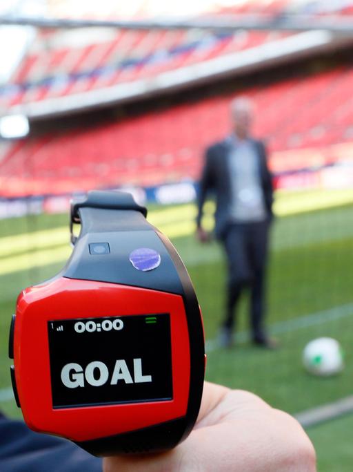 Eine Art Armbanduhr mit der Aufschrift "Goal", im Hintergrund ein Ball in einem Tor in einem Fußballstadion.
